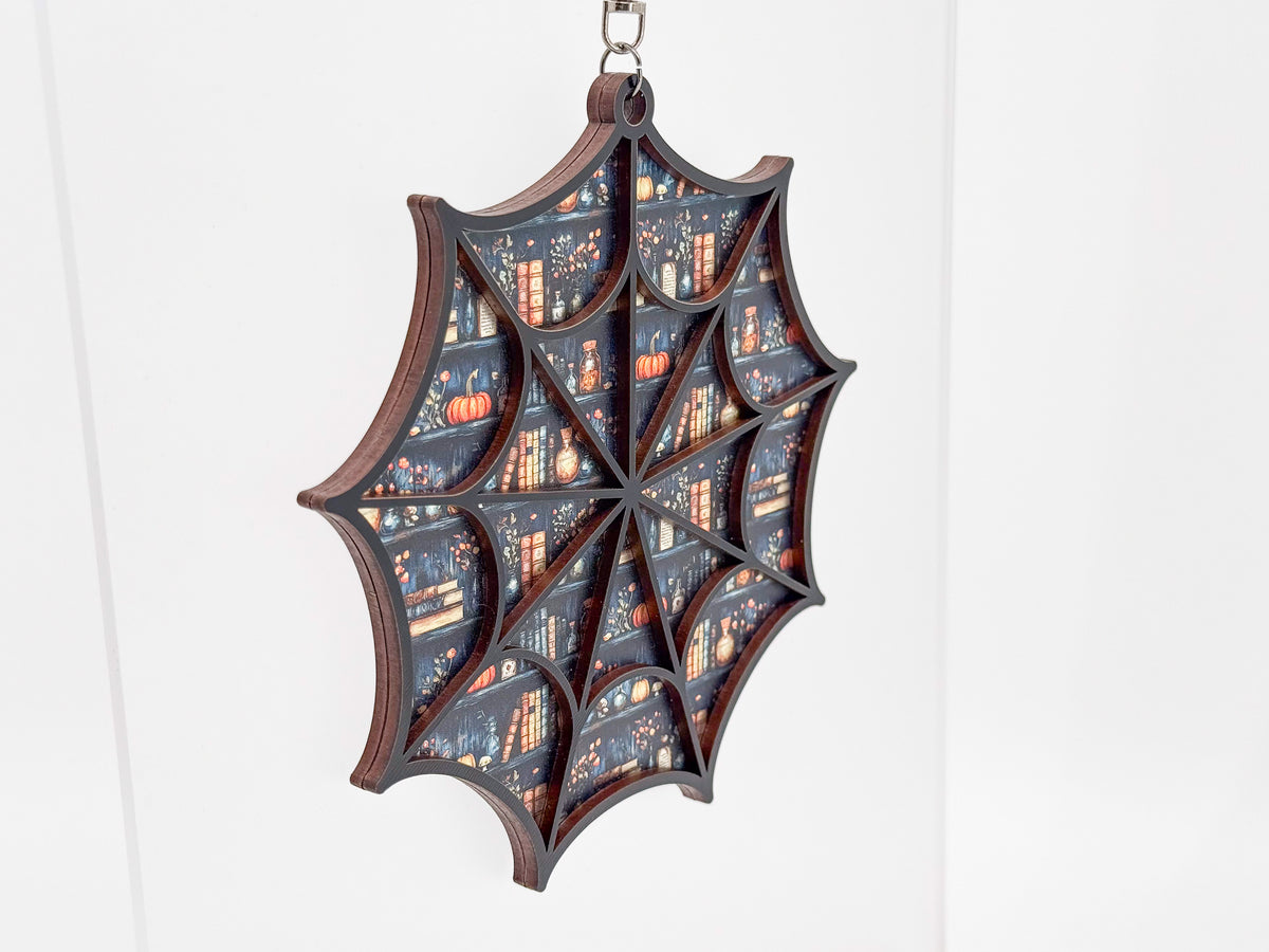 Angled view of the Fall Bookshelves spiderweb suncatcher, showing layered wood edges and a pattern of books, pumpkins, and bottles.