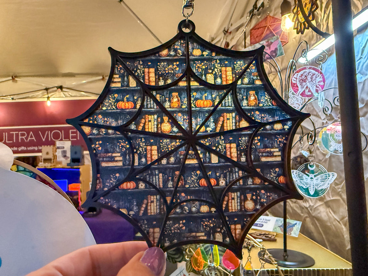 Fall Bookshelves spiderweb suncatcher hanging at a display booth, with a hand holding its lower edge.
