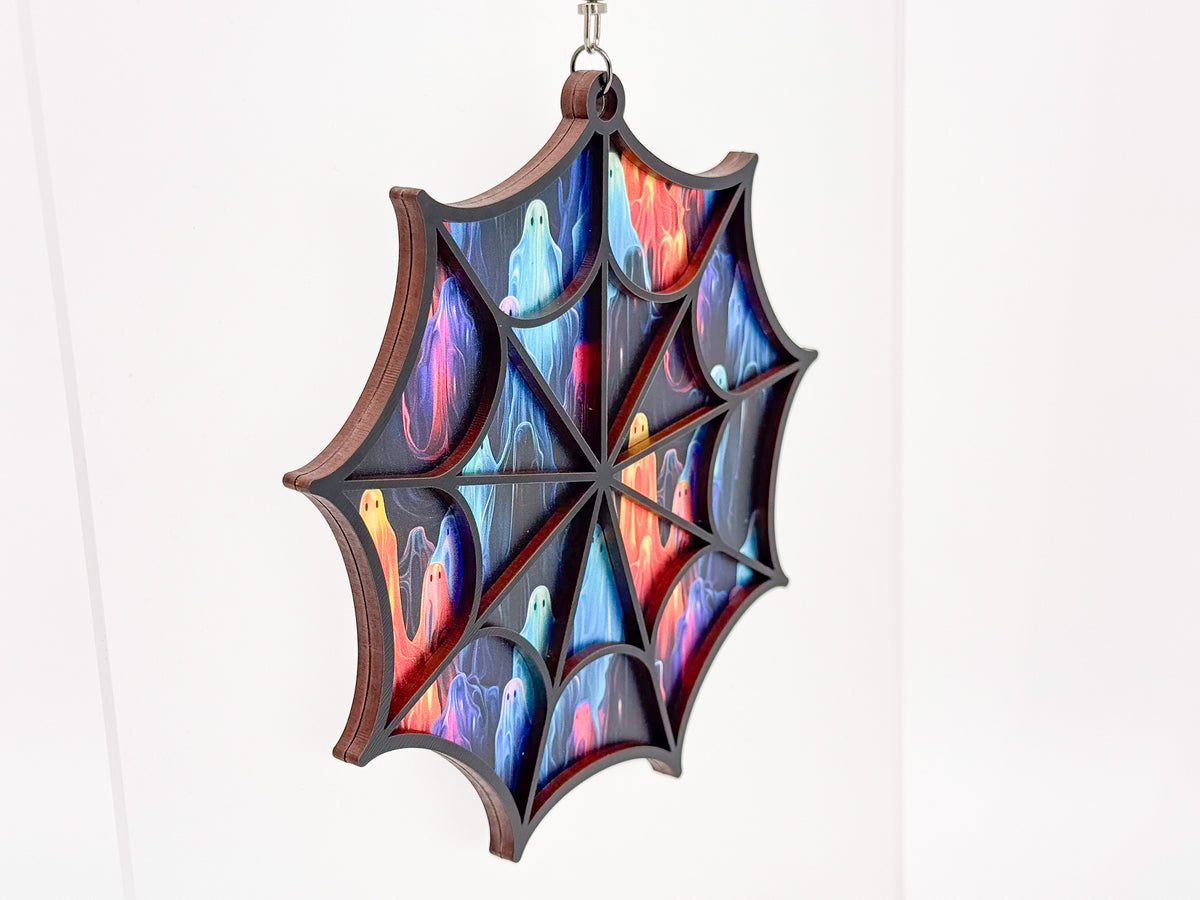 Angled view of the Ghosts spiderweb suncatcher, showing layered wood edges and colorful ghosts behind the black web frame.