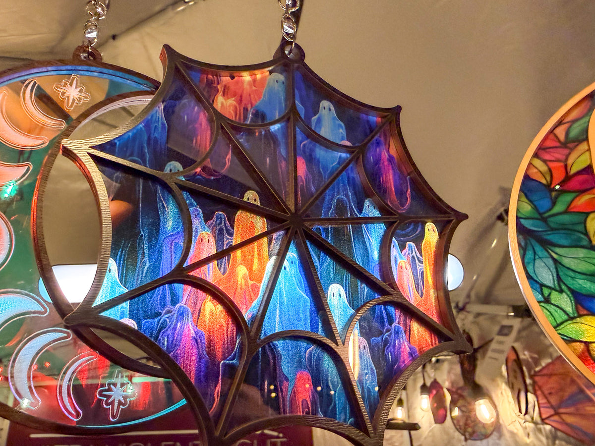 Colorful ghost-pattern spiderweb suncatcher illuminated in a display beside other hanging suncatchers.