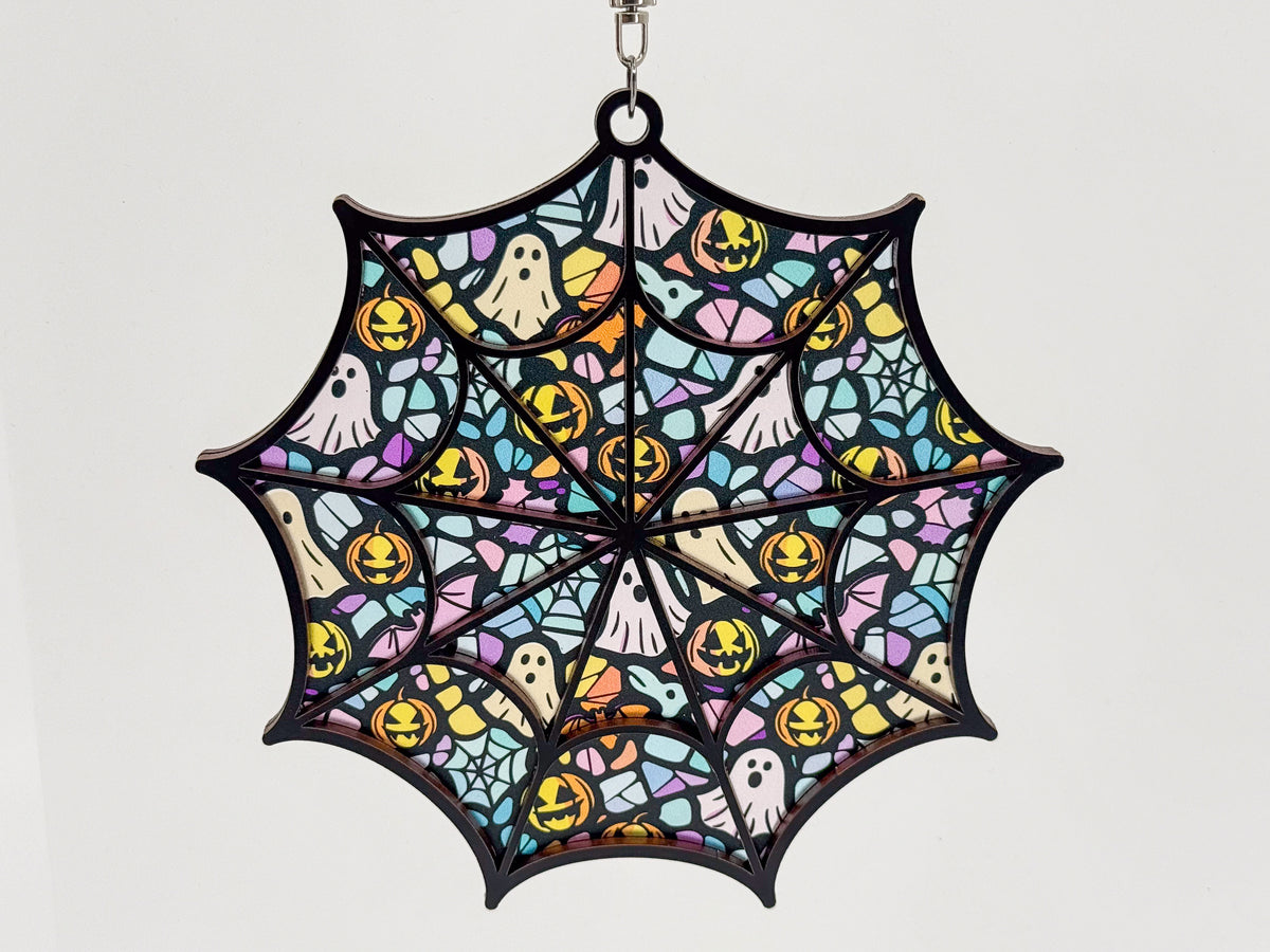 Front view of a spiderweb suncatcher with pastel ghosts, yellow jack-o’-lanterns, and small webs behind a black wood frame.