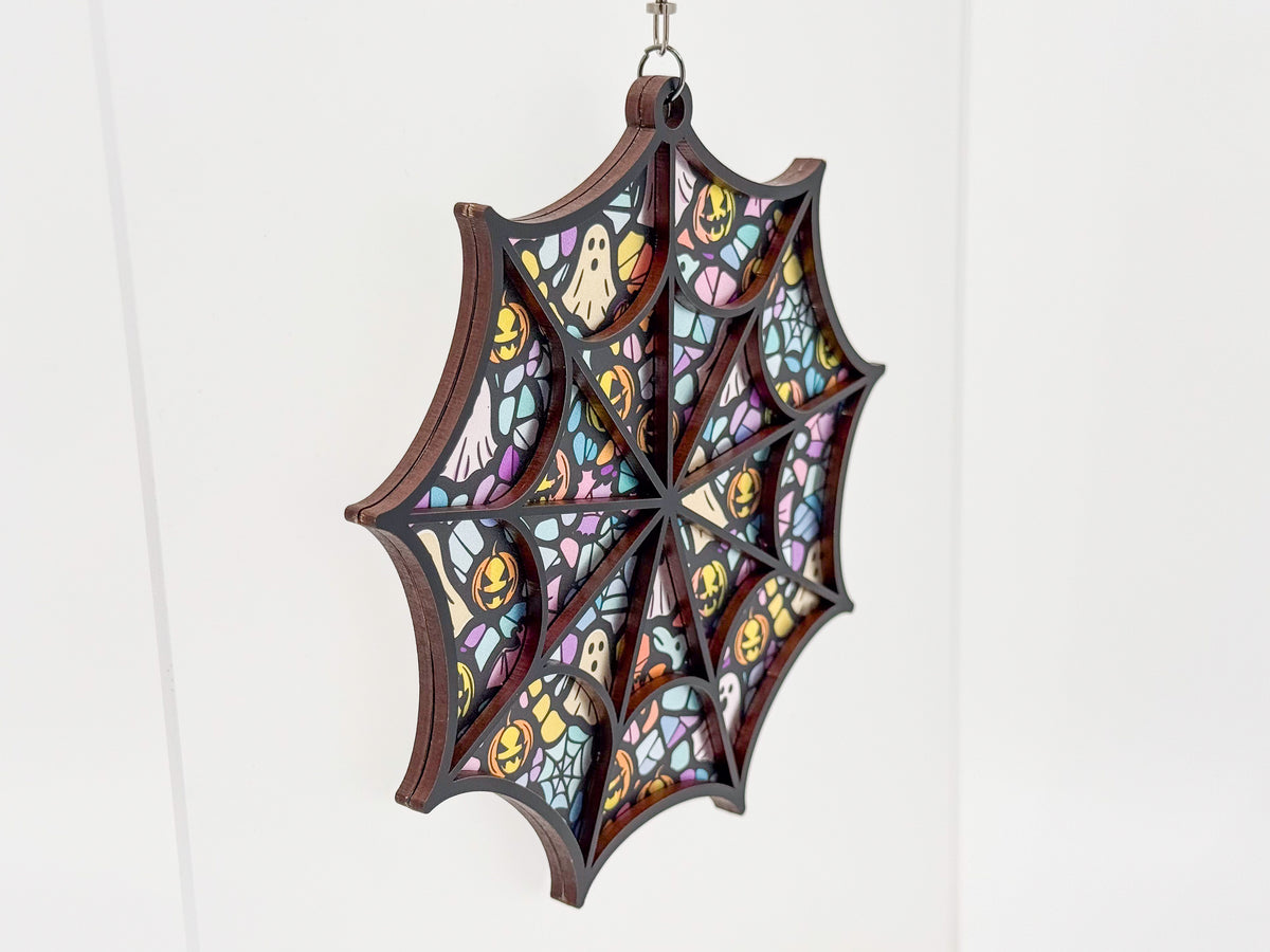 Angled view of the Whimsical spiderweb suncatcher, showing layered wood edges and a pastel ghost and jack-o’-lantern pattern.