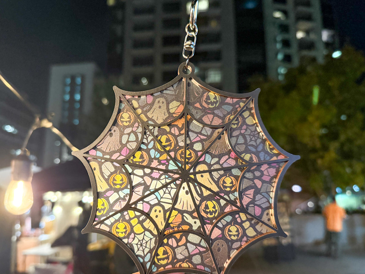 Whimsical spiderweb suncatcher hanging outdoors at night, with pastel ghosts and yellow jack-o’-lanterns illuminated beside string lights.