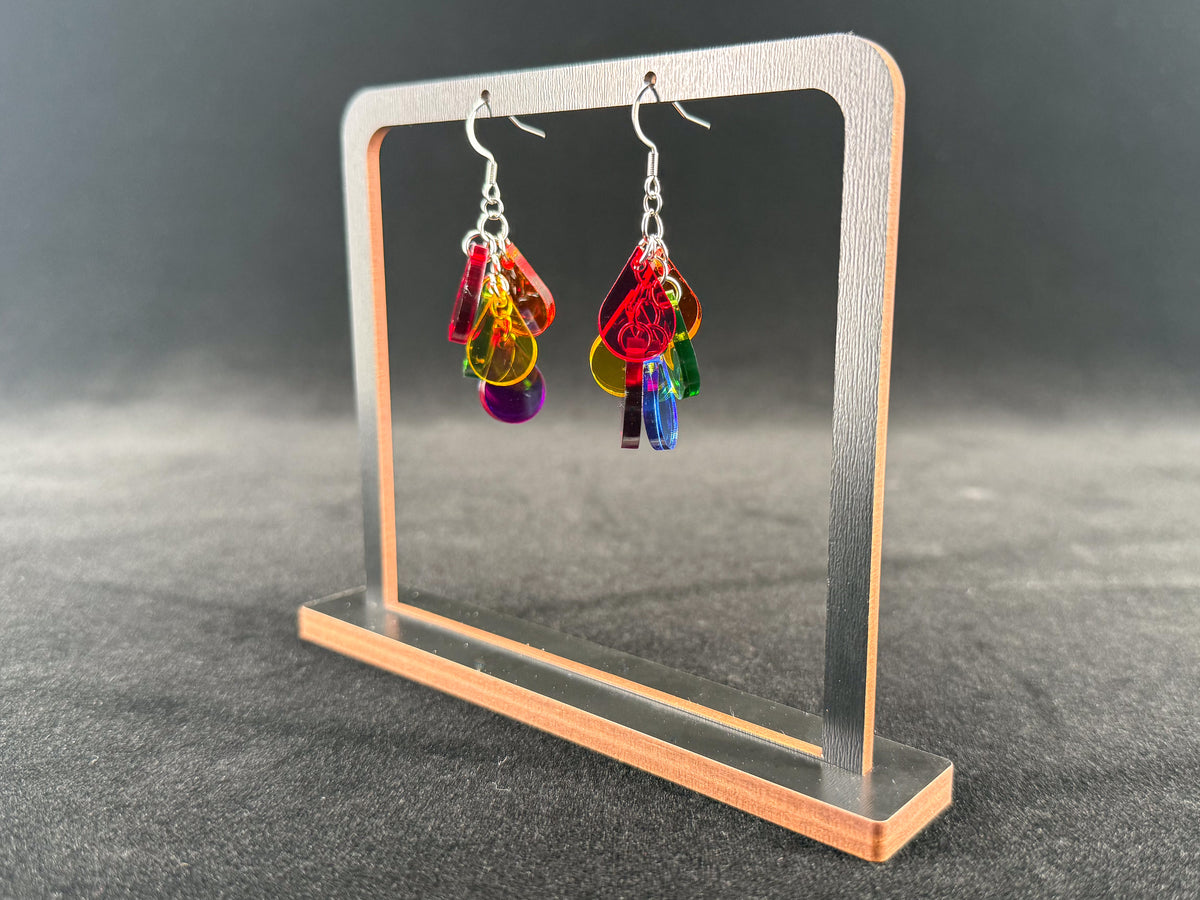 A pair of colorful dangling earrings displayed on a clear acrylic stand against a dark gray background. The earrings feature multiple translucent dangling charms in red, orange, yellow, green, blue, and purple with silver fishhook wires.