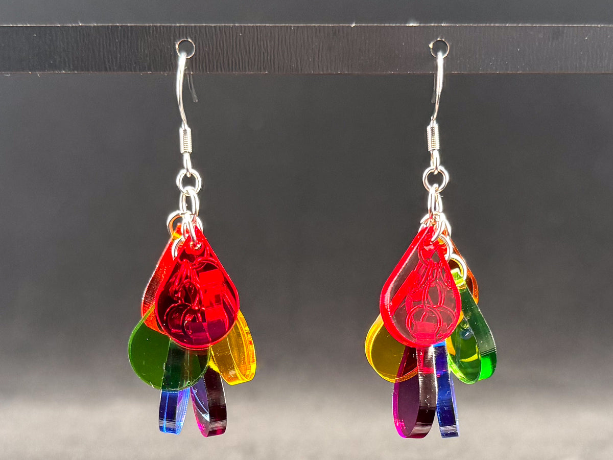 A pair of colorful acrylic dangle earrings hanging from silver fishhook wires. The earrings feature multiple translucent teardrop-shaped pieces in red, green, yellow, blue, purple, and pink, with some pieces cut into thin strips at the bottom, set against a dark gray background.