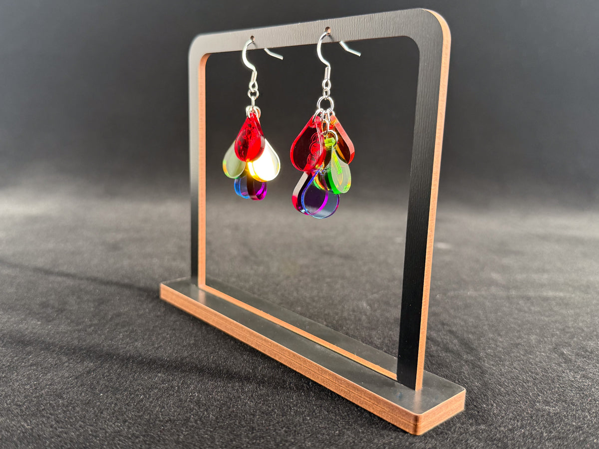 A pair of colorful dangling earrings displayed on a small metal stand against a dark gray background. The earrings have silver fishhook wires and multiple translucent teardrop charms in red, green, blue, yellow, and white.