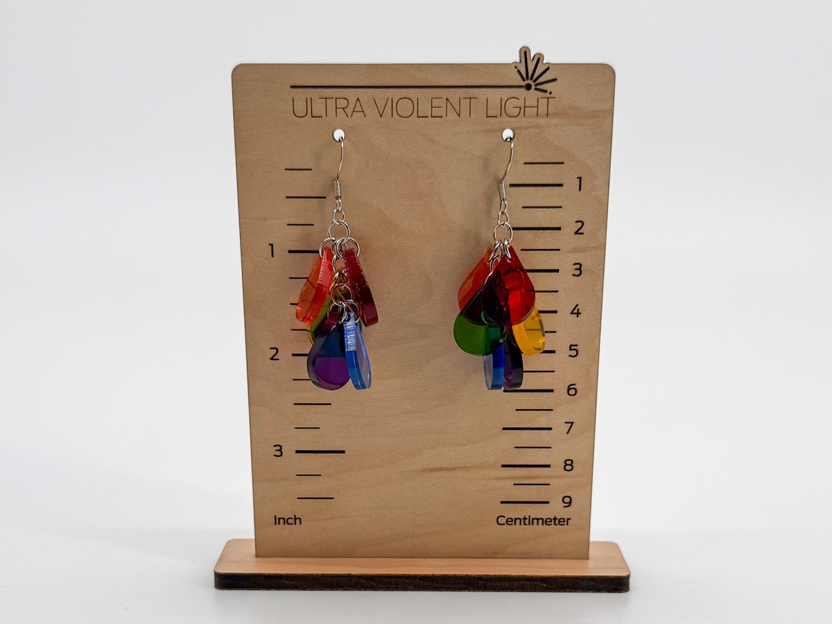 A pair of colorful dangling earrings displayed on a wooden height-measuring stand labeled 'ULTRA VIOLENT LIGHT.' The earrings have translucent rainbow-colored petal-like charms with silver hooks.