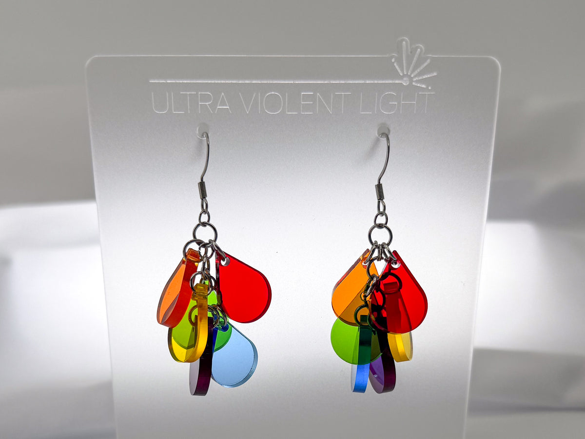 A pair of colorful acrylic dangle earrings displayed on a clear acrylic stand. The earrings feature multiple translucent color panels in red, orange, yellow, green, blue, and purple, hanging from silver fishhook wires.
