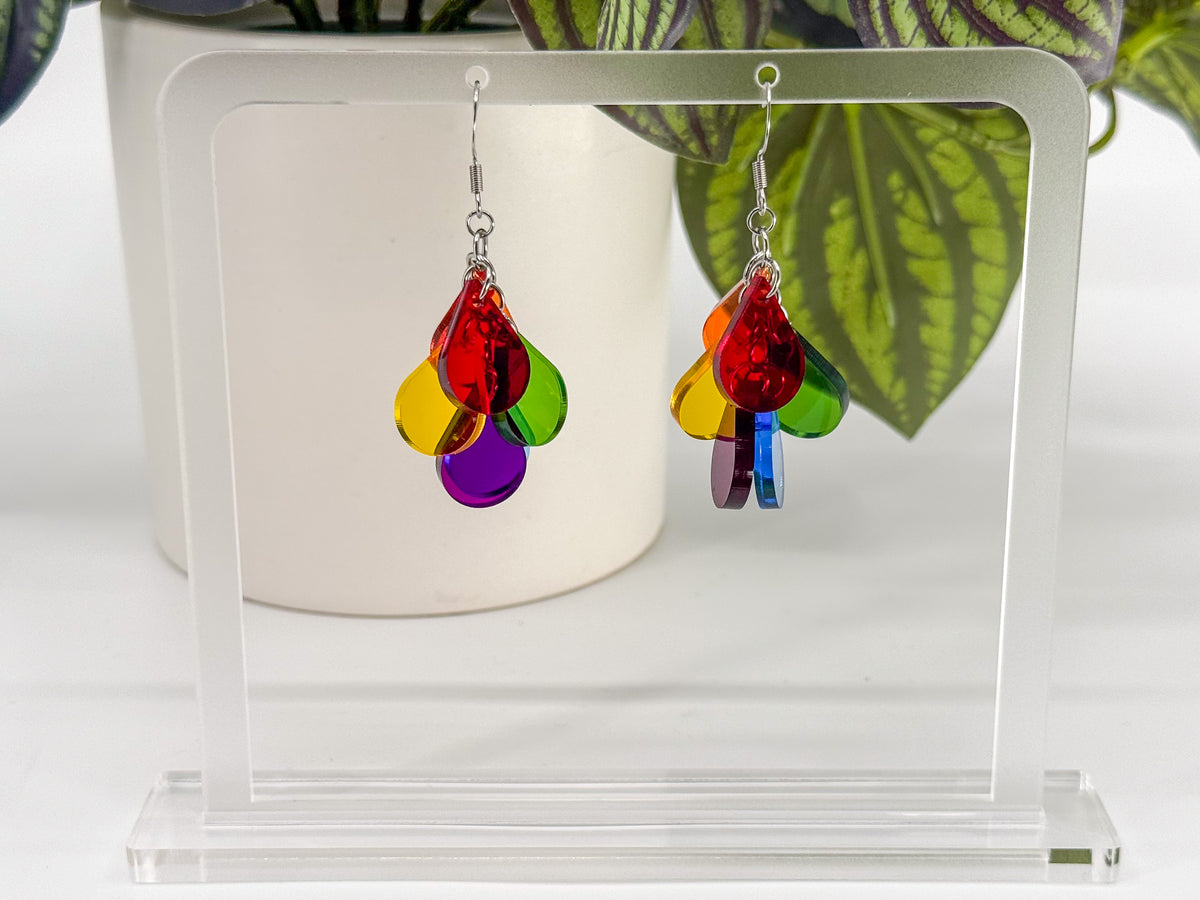 Rainbow-colored dangling earrings with layered translucent teardrop shapes in red, orange, yellow, green, blue, and purple, displayed on a clear acrylic stand in front of a white planter with green leaves.