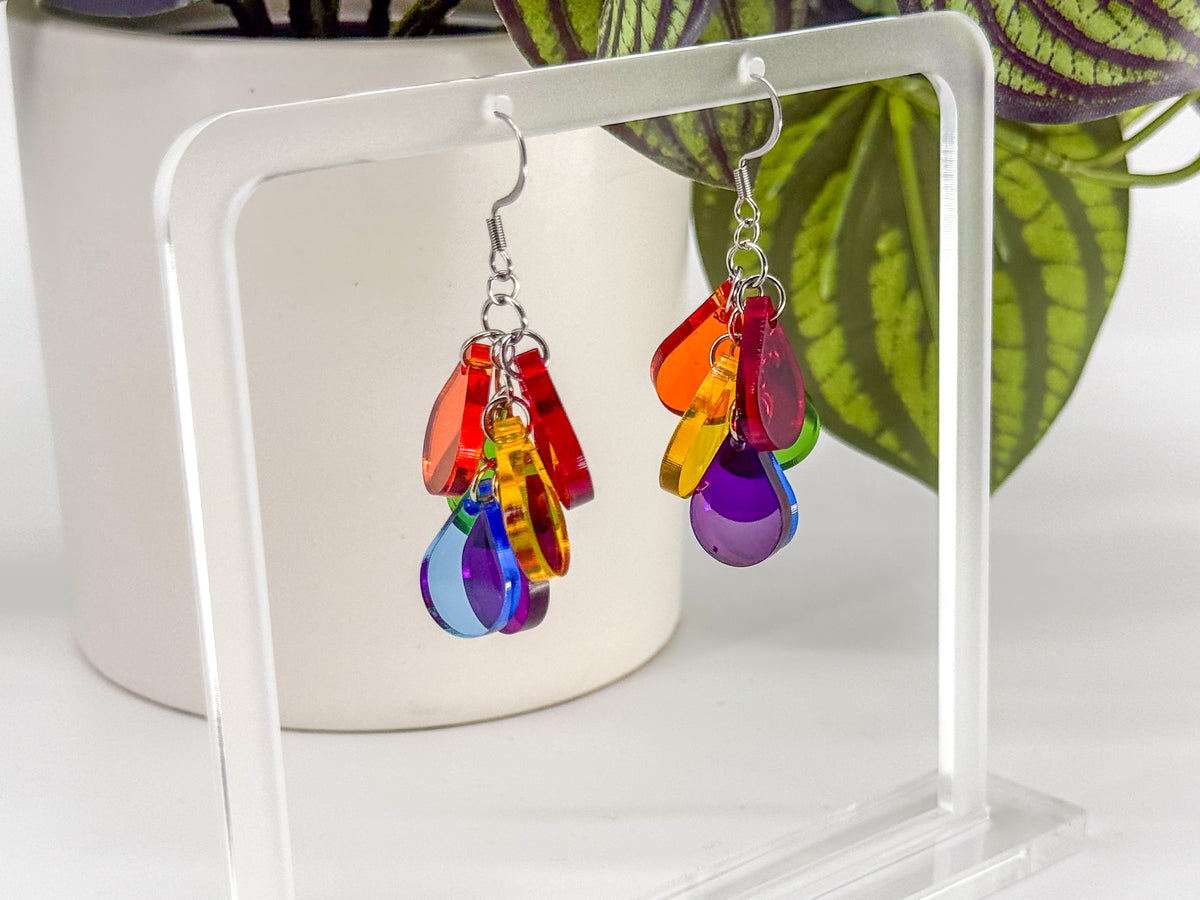 A pair of dangling earrings with translucent rainbow-colored teardrop charms on silver hooks, displayed on a clear acrylic stand in front of a white pot and green plant leaves.