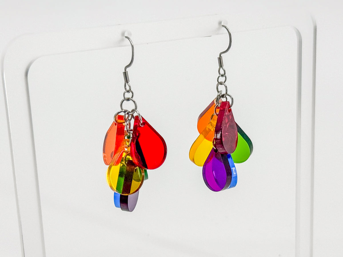 A pair of rainbow-colored dangling earrings with translucent heart-shaped charms in red, orange, yellow, green, blue, and purple, hanging from silver fishhook wires against a white background.