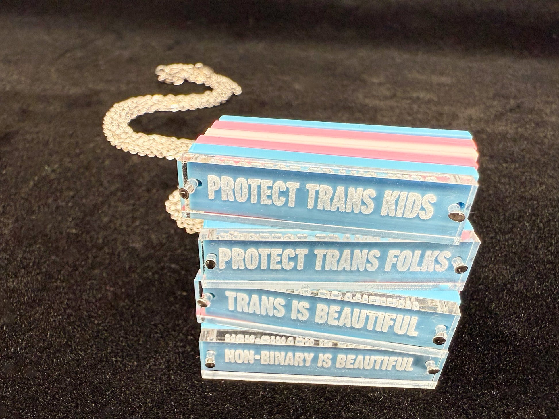 Four necklaces stacked, each reading different phrases: Protect trans kids, Protect trans folks, Trans is beautiful, Non-binary is beautiful