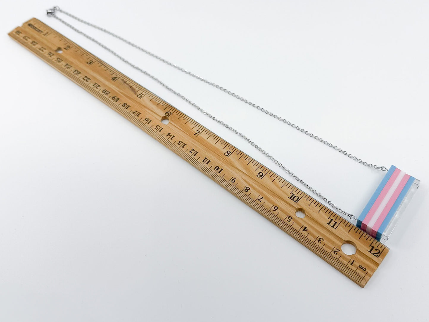 A trans pride flag necklace shown roughly the size of an adjacent foot-long ruler.