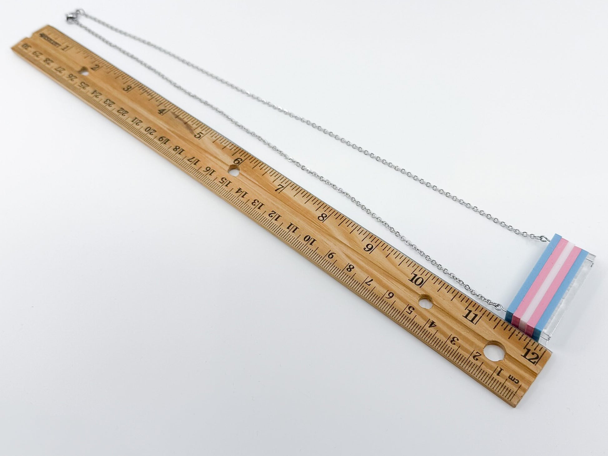 A trans pride flag necklace shown roughly the size of an adjacent foot-long ruler.