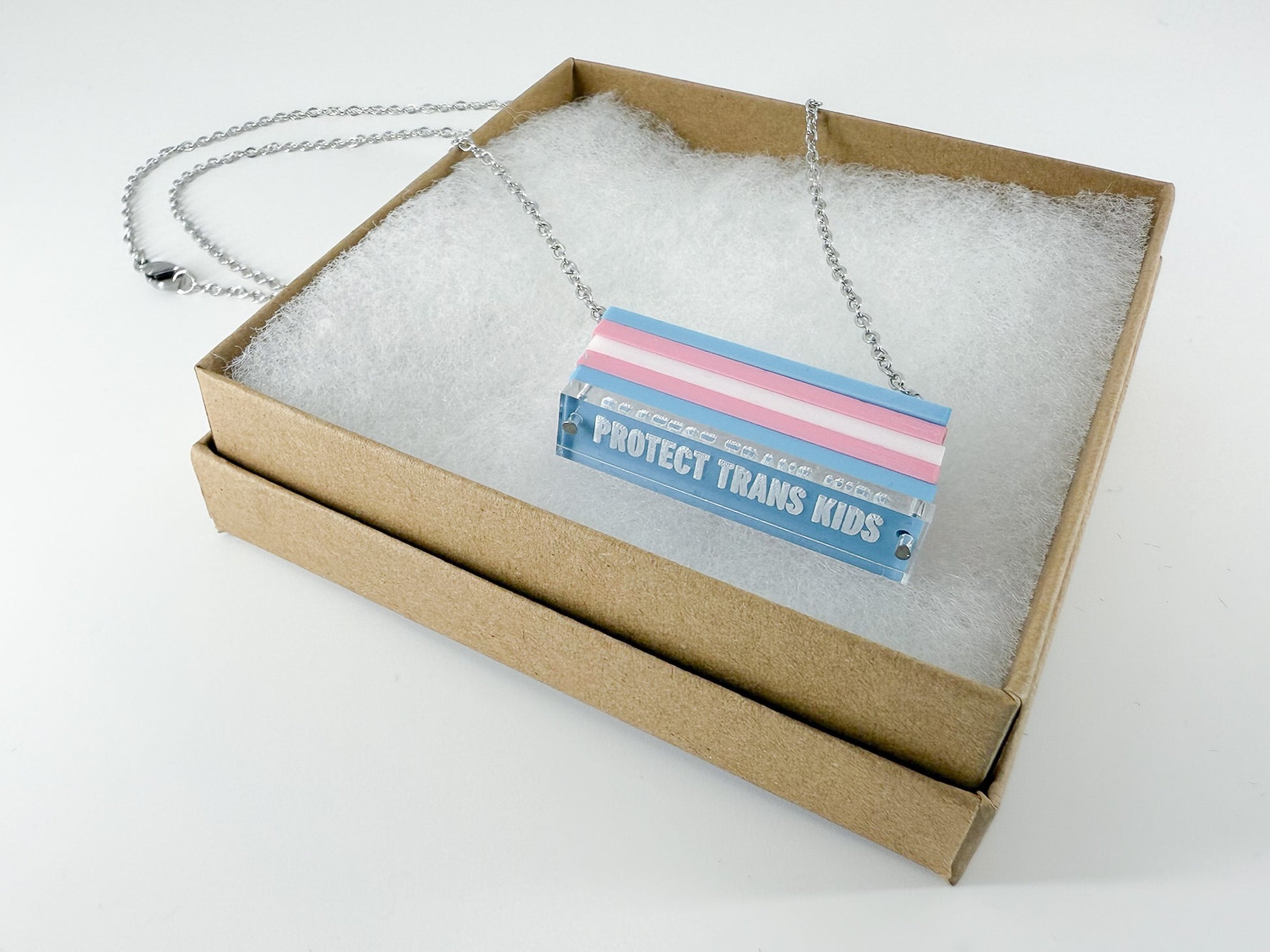 A trans pride flag necklace, with the words "protect trans kids" engraved at the bottom, sitting in a Kraft paper gift box.