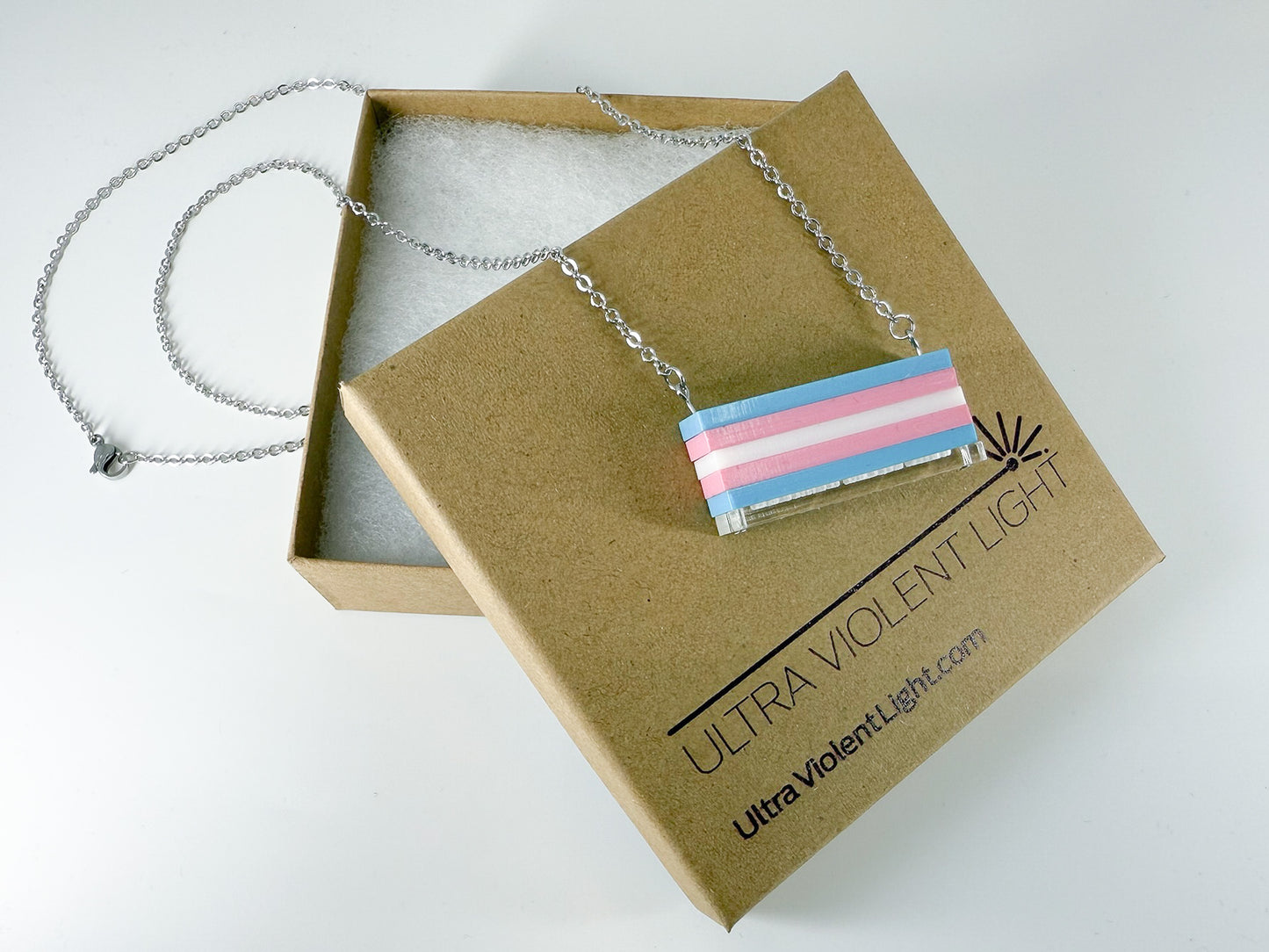 A trans pride flag necklace draped over a gift jewelry box.