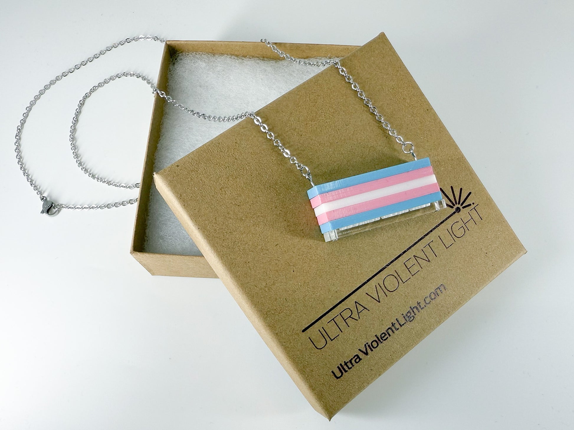 A trans pride flag necklace draped over a gift jewelry box.