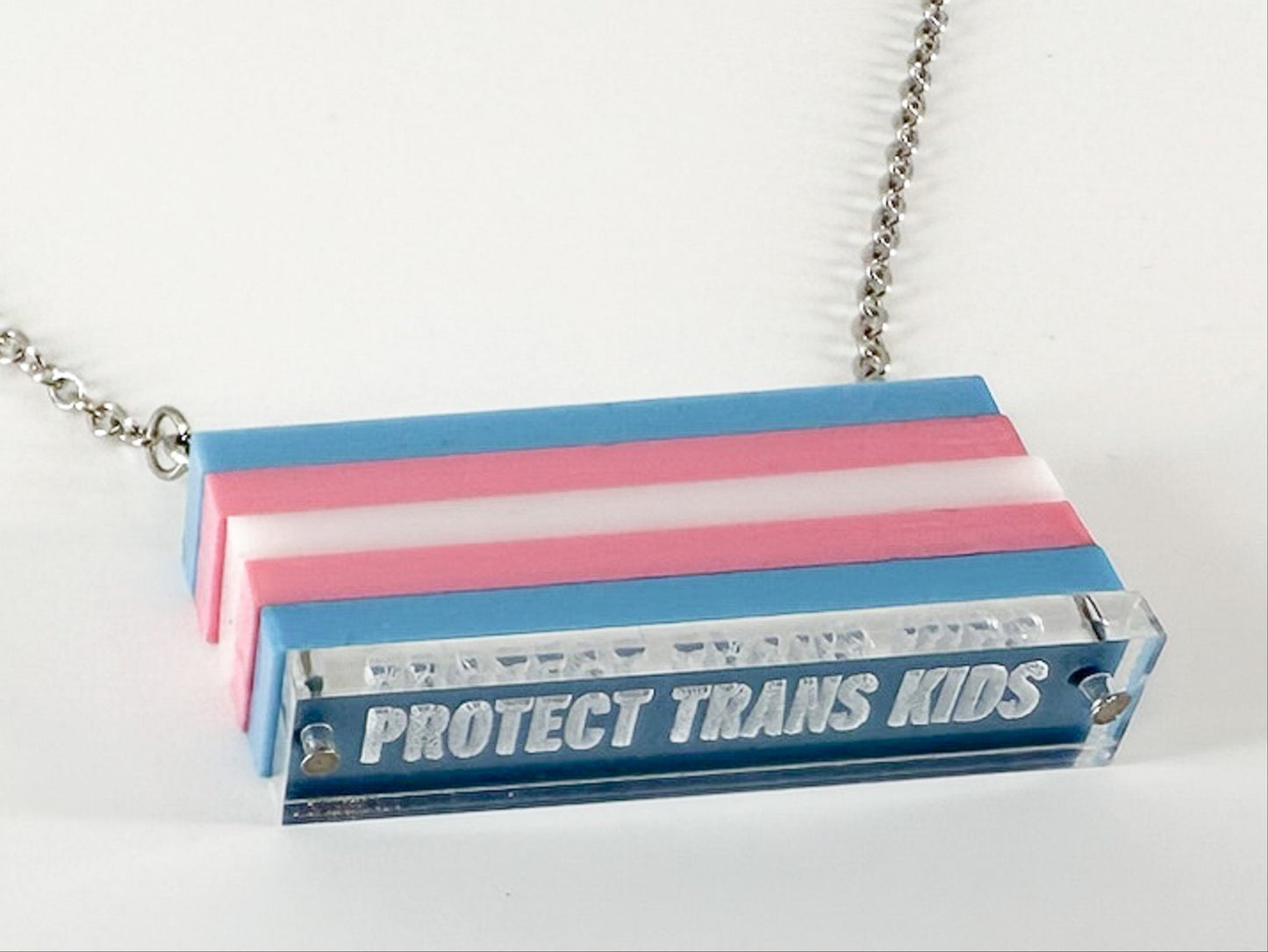 A close-up of a trans pride flag necklace, with the words "protect trans kids" engraved at the bottom.
