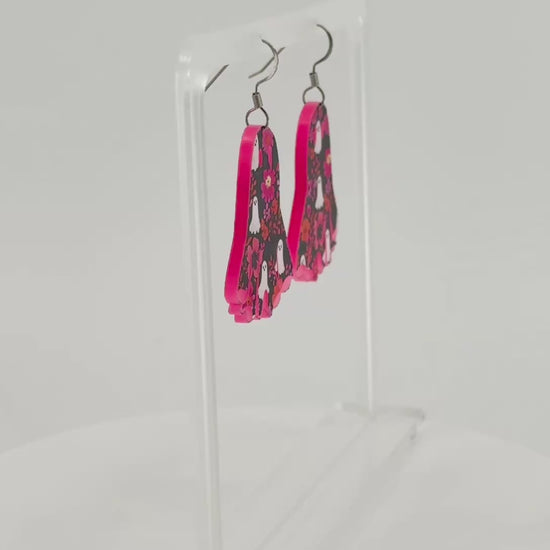 A pair of ghost-shaped dangle earrings with a pink, black, and white floral ghost pattern, displayed on a clear acrylic stand.