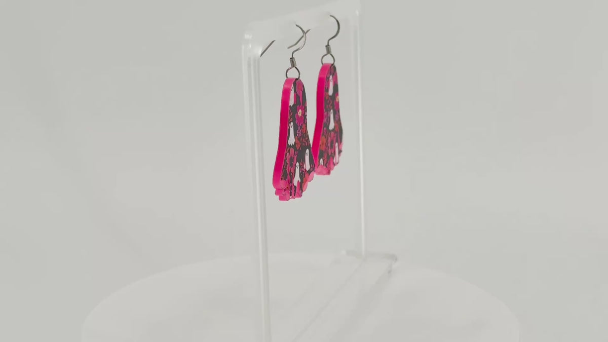 A pair of ghost-shaped dangle earrings with a pink, black, and white floral ghost pattern, displayed on a clear acrylic stand.