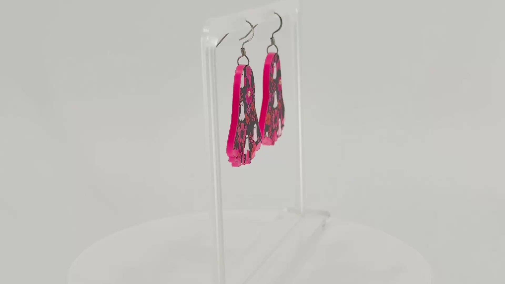 A pair of ghost-shaped dangle earrings with a pink, black, and white floral ghost pattern, displayed on a clear acrylic stand.