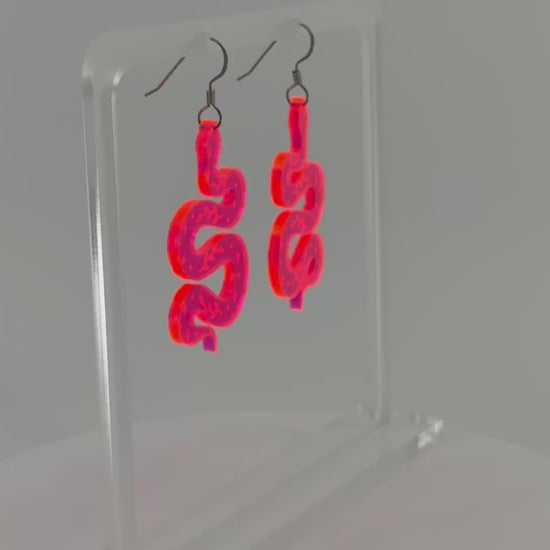 A pair of fluorescent pink snake-shaped dangle earrings displayed on a clear acrylic stand against a white background, spinning to show all sides.