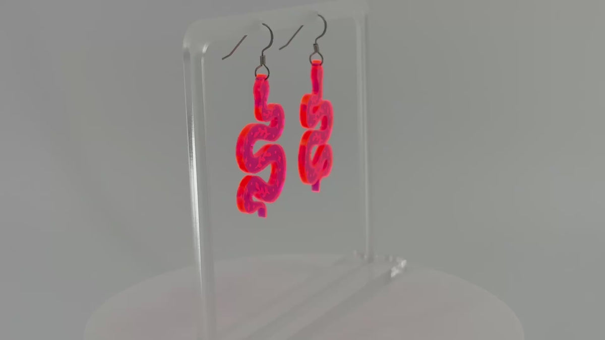 A pair of fluorescent pink snake-shaped dangle earrings displayed on a clear acrylic stand against a white background, spinning to show all sides.