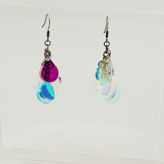 A pair of dangling earrings displayed on a clear acrylic stand. Each earring has a silver fishhook ear wire with a cluster of dangling iridescent teardrop shaped charms. The stand is being rotated for the video.