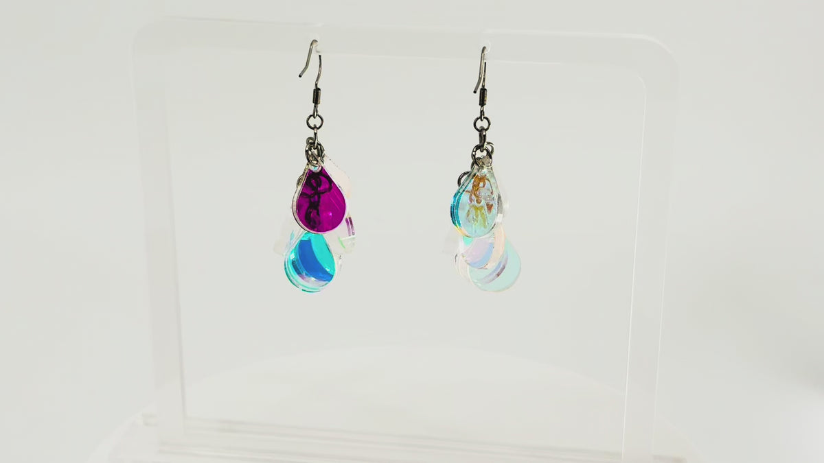 A pair of dangling earrings displayed on a clear acrylic stand. Each earring has a silver fishhook ear wire with a cluster of dangling iridescent teardrop shaped charms. The stand is being rotated for the video.