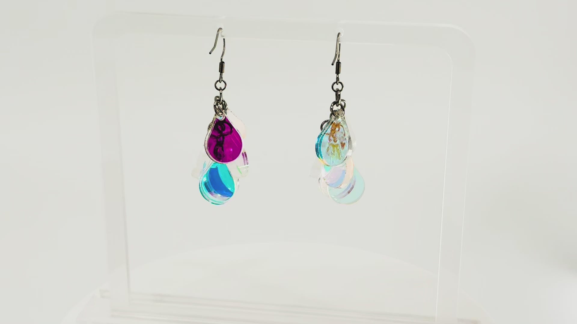 A pair of dangling earrings displayed on a clear acrylic stand. Each earring has a silver fishhook ear wire with a cluster of dangling iridescent teardrop shaped charms. The stand is being rotated for the video.