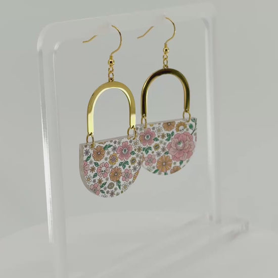 A pair of dangling earrings displayed on a clear acrylic stand. Each earring has a gold-tone arch top and a curved lower section covered in a dense multicolor floral pattern with pink, orange, yellow, green, and blue tones, featuring hidden skulls and eyes. The earrings and stand rotate in the video.