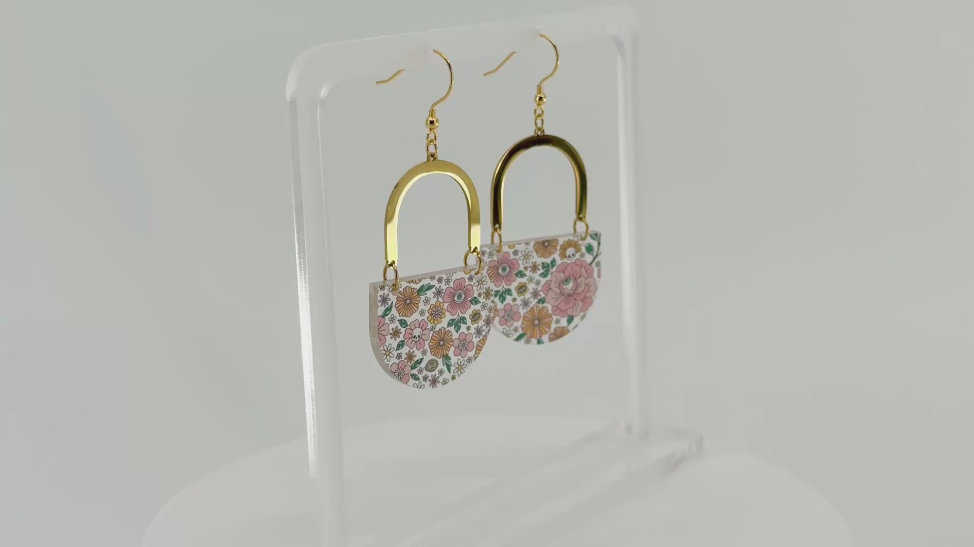 A pair of dangling earrings displayed on a clear acrylic stand. Each earring has a gold-tone arch top and a curved lower section covered in a dense multicolor floral pattern with pink, orange, yellow, green, and blue tones, featuring hidden skulls and eyes. The earrings and stand rotate in the video.