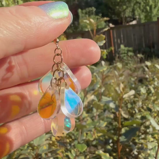 A hand holds a pair of dangling earrings displayed in the sunshine in front of some plants. Each earring has a gold fishhook ear wire with a cluster of dangling iridescent teardrop shaped charms.