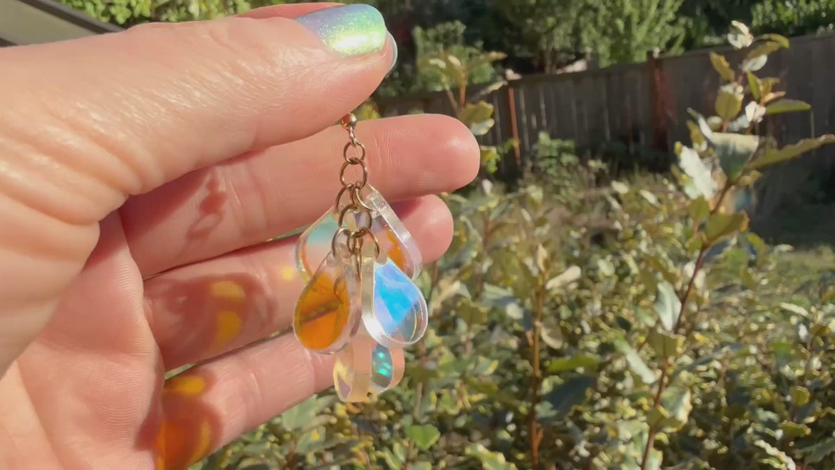A hand holds a pair of dangling earrings displayed in the sunshine in front of some plants. Each earring has a gold fishhook ear wire with a cluster of dangling iridescent teardrop shaped charms.