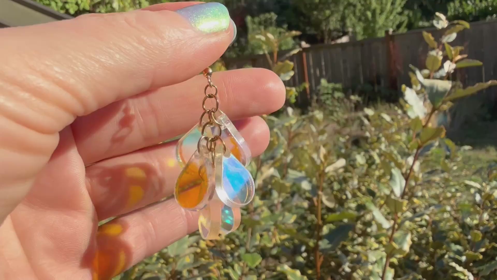 A hand holds a pair of dangling earrings displayed in the sunshine in front of some plants. Each earring has a gold fishhook ear wire with a cluster of dangling iridescent teardrop shaped charms.