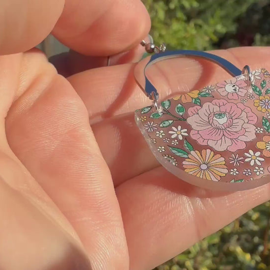 A hand holds a dangling earring with a silver-tone arch top and a curved lower section covered in a dense multicolor floral pattern with pink, orange, yellow, green, and blue tones, featuring hidden skulls and eyes. The hand and earring are outside, in front of green plants, and the hand moves the earring around to show motion and transparency.