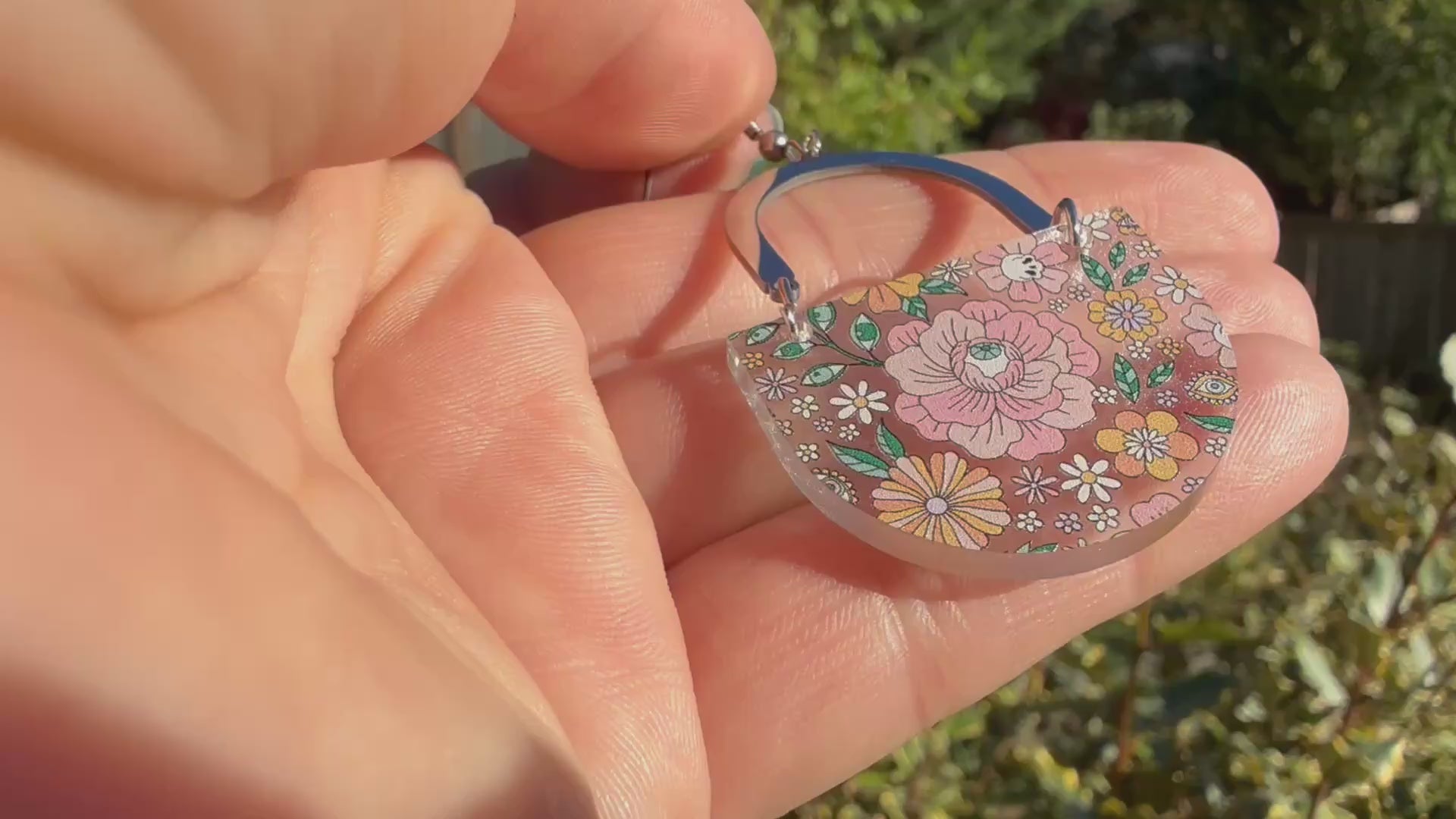 A hand holds a dangling earring with a silver-tone arch top and a curved lower section covered in a dense multicolor floral pattern with pink, orange, yellow, green, and blue tones, featuring hidden skulls and eyes. The hand and earring are outside, in front of green plants, and the hand moves the earring around to show motion and transparency.