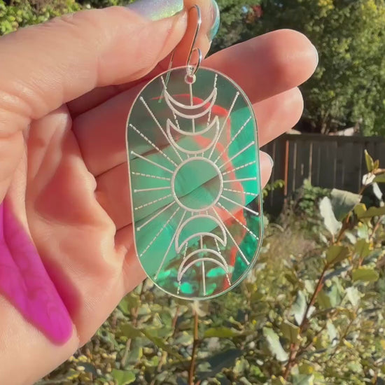 A hand holds a single oval drop earring with an engraved crescent moon and sun design, featuring a blue-to-purple-to pink iridescent background, displayed moving with different angles of sunlight shining through.