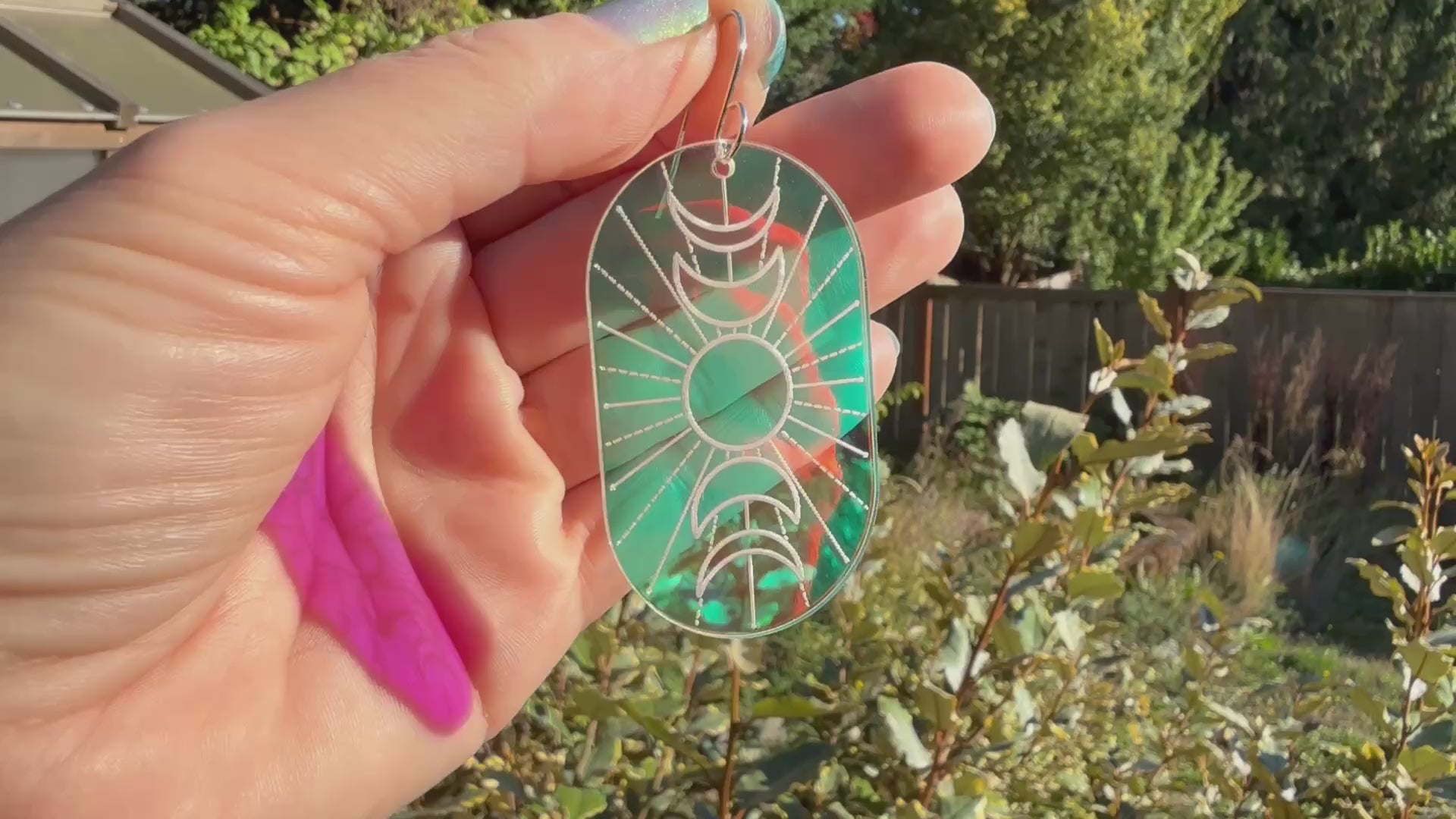 A hand holds a single oval drop earring with an engraved crescent moon and sun design, featuring a blue-to-purple-to pink iridescent background, displayed moving with different angles of sunlight shining through.