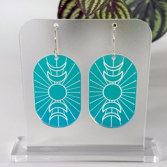A pair of oval drop earrings with a engraved crescent moon and sun design, featuring a blue-to-purple gradient background, hanging on a clear acrylic display stand. The stand and earrings rotate in the video, displaying a range of colors as the iridescence changes with the shift of light.