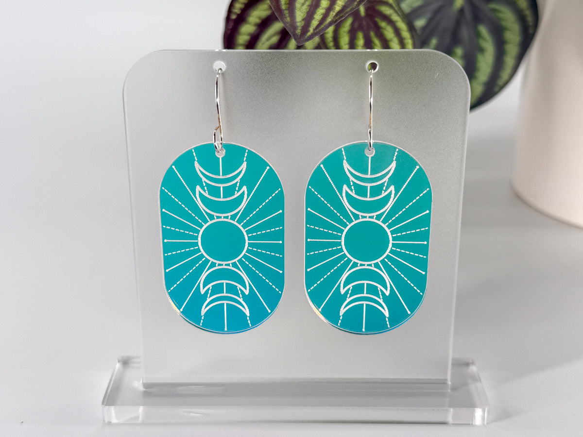 A pair of oval drop earrings with a engraved crescent moon and sun design, featuring a blue-to-purple gradient background, hanging on a clear acrylic display stand. The stand and earrings rotate in the video, displaying a range of colors as the iridescence changes with the shift of light.