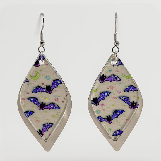 A pair of long, smooth rhombus-shaped dangle earrings with a beige background and a pattern of purple, blue, and green bats and moons, displayed on a clear acrylic stand against a white background. The earrings and stand are rotating in the video.