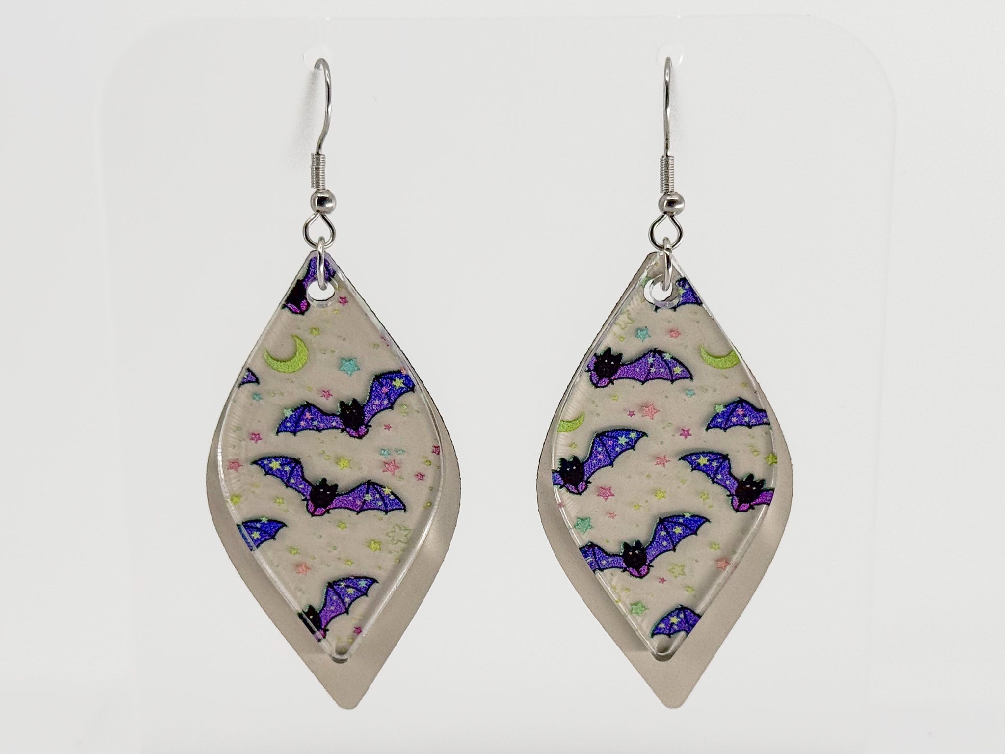 A pair of long, smooth rhombus-shaped dangle earrings with a beige background and a pattern of purple, blue, and green bats and moons, displayed on a clear acrylic stand against a white background. The earrings and stand are rotating in the video.