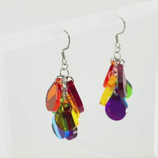 A pair of colorful acrylic dangle earrings displayed on a clear acrylic stand. The earrings feature multiple translucent color panels in red, orange, yellow, green, blue, and purple, hanging from silver fishhook wires. The stand is rotating in the picture, showing all sides of the earrings.