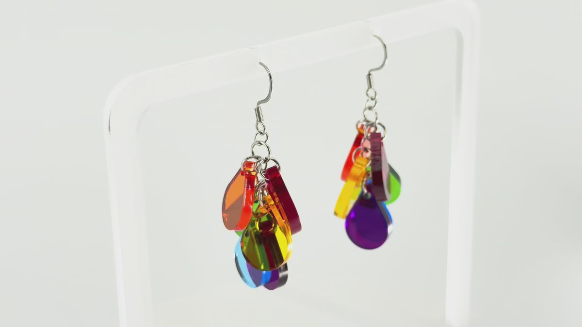 A pair of colorful acrylic dangle earrings displayed on a clear acrylic stand. The earrings feature multiple translucent color panels in red, orange, yellow, green, blue, and purple, hanging from silver fishhook wires. The stand is rotating in the picture, showing all sides of the earrings.