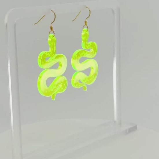 A pair of fluorescent green snake-shaped dangle earrings displayed on a clear acrylic stand against a white background, spinning to show all sides.