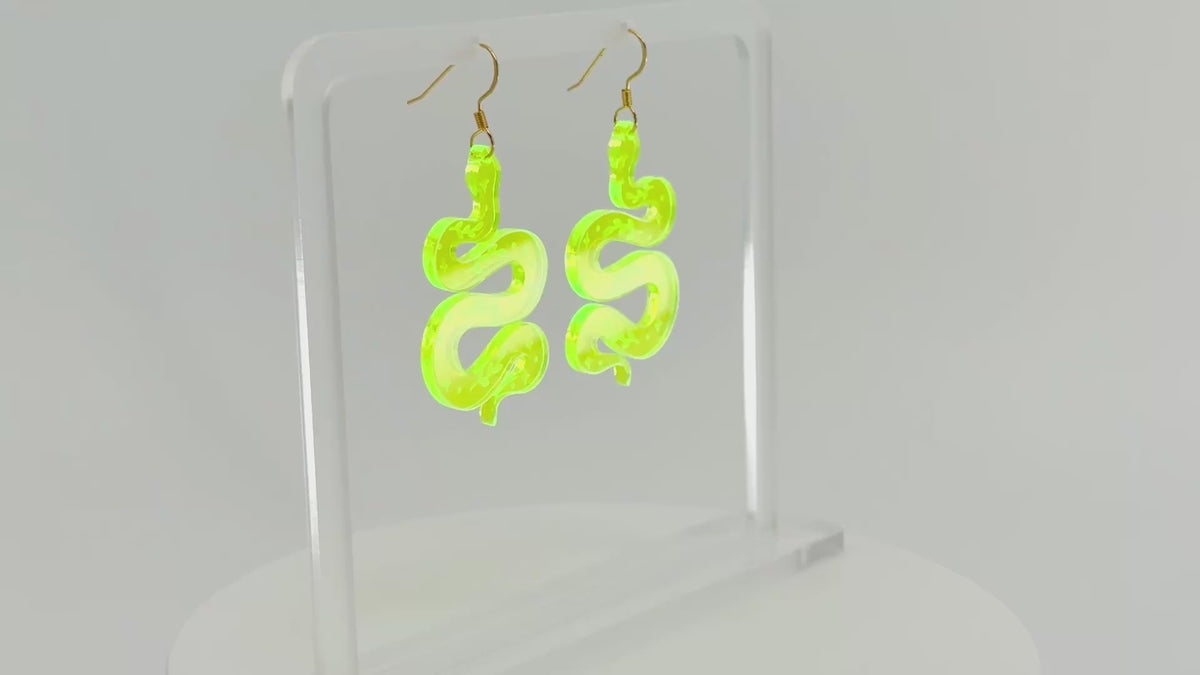 A pair of fluorescent green snake-shaped dangle earrings displayed on a clear acrylic stand against a white background, spinning to show all sides.