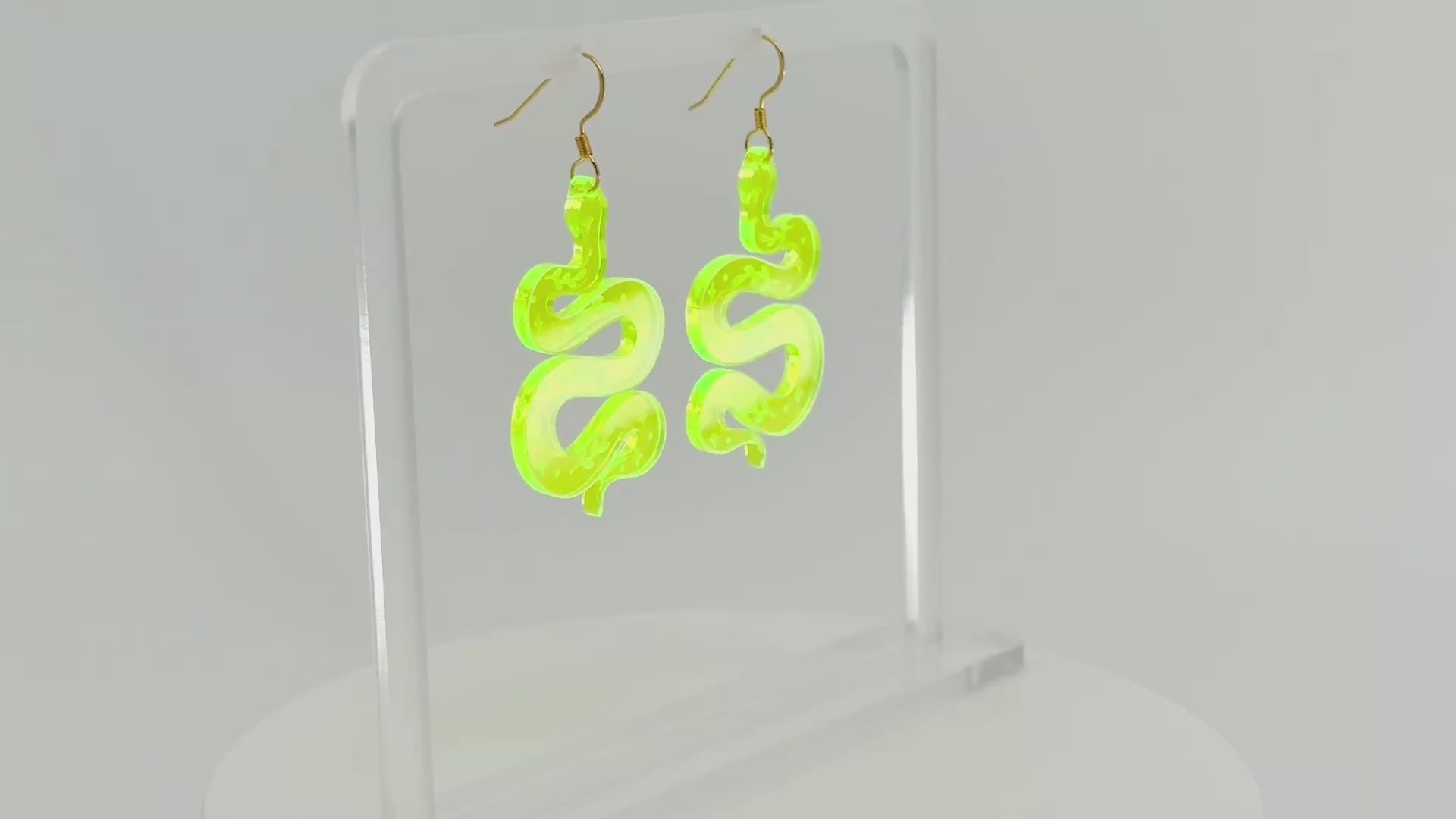 A pair of fluorescent green snake-shaped dangle earrings displayed on a clear acrylic stand against a white background, spinning to show all sides.