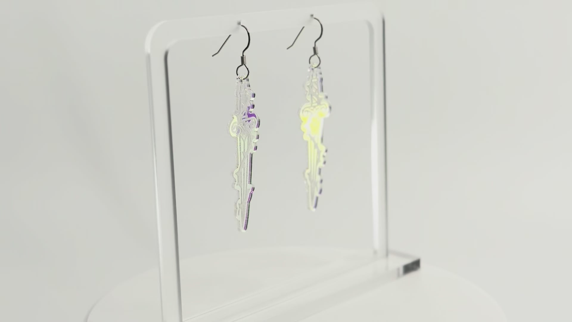 Iridescent sword earrings are displayed on a clear acrylic stand. The stand and earrings are rotating to see all sides.