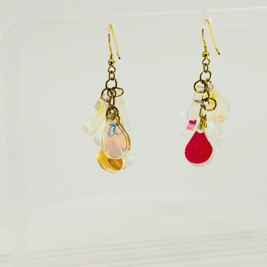 A pair of dangling earrings displayed on a clear acrylic stand. Each earring has a gold fishhook ear wire with a cluster of dangling iridescent teardrop shaped charms. The stand is being rotated for the video.